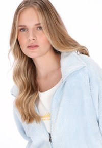 Młoda kobieta o długich blond włosach nosi jasnoniebieską bluzę polarową na zamek na białą koszulę, patrzy prosto przed siebie z neutralnym wyrazem twarzy.