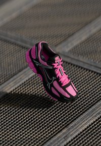 Ružová a čierna bežecká obuv Nike Vomero s priezračným dizajnom položená na kovovej mriežke vonku.