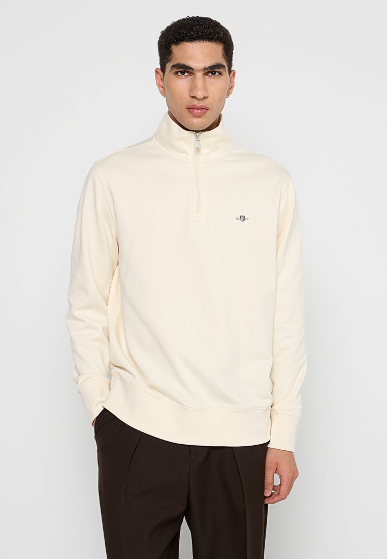 Junger Mann in einem cremefarbenen Halbzip-Pullover und dunklen Hosen, der mit den Händen in den Taschen vor einem schlichten hellen Hintergrund steht.