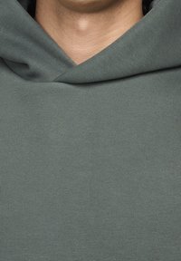 Mikina s kapucí v tlumené zelené barvě, vyrobená z hladkého materiálu a s volným střihem, která se vyznačuje minimalistickým designem bez zdobení.