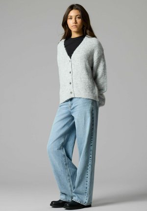 Jauna moteris dėvi šviesiai pilką pūkuotą kardiganą ant juodo viršaus, plačias mėlynas džinsines kelnes ir juodus batus, stovinti priešais paprastą pilką foną.