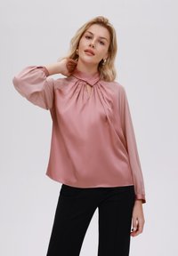 Blusa di raso rosa pallido con design a pieghe, morbide maniche a sbuffo e dettaglio del collo a fessura. Abbinata a pantaloni neri.