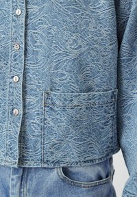 Camicia di denim azzurro chiaro con un motivo testurizzato a spirale, dotata di chiusure a bottoni e una tasca applicata sul lato sinistro anteriore.