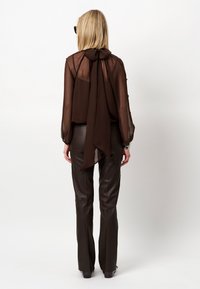 Bruine doorschijnende blouse met een strik aan de achterkant en lange mouwen, gecombineerd met donkere lerenachtige broek. Verspreide knopen sieren de mouwen.