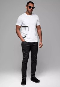 Hvid t-shirt med sorte geometriske detaljer, parret med sorte slim-fit bukser og sorte sneakers. Modellen bærer mørke solbriller.