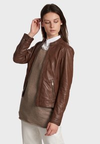 Bruine leren jas met een gladde textuur, ritssluiting aan de voorkant en zijzakken. Heeft een ronde kraag en een getailleerd ontwerp, gedragen over een lichte trui.