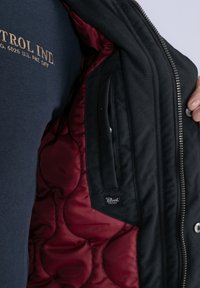 Giacca nera con una fodera trapuntata bordeaux, dotata di chiusura con zip, tasca laterale e un piccolo etichetta del brand all'interno.