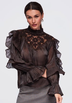 Femme portant un chemisier brun foncé transparent avec un empiècement en dentelle florale et des manches à volants, associé à une jupe à motif pied-de-poule.
