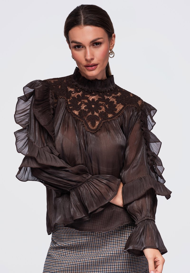 Femme portant un chemisier brun foncé transparent avec un empiècement en dentelle florale et des manches à volants, associé à une jupe à motif pied-de-poule.