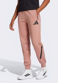 Άτομο που φοράει ανοιχτό ροζ Adidas κολάν με μαύρο λογότυπο και λευκά Adidas sneakers, στέκεται μπροστά από απλό φόντο.