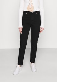 Jeans de denim preto com um corte slim, design de cinco bolsos e discretos detalhes em cobre. Combinados com uma blusa de manga longa branca canelada e ténis brancos.