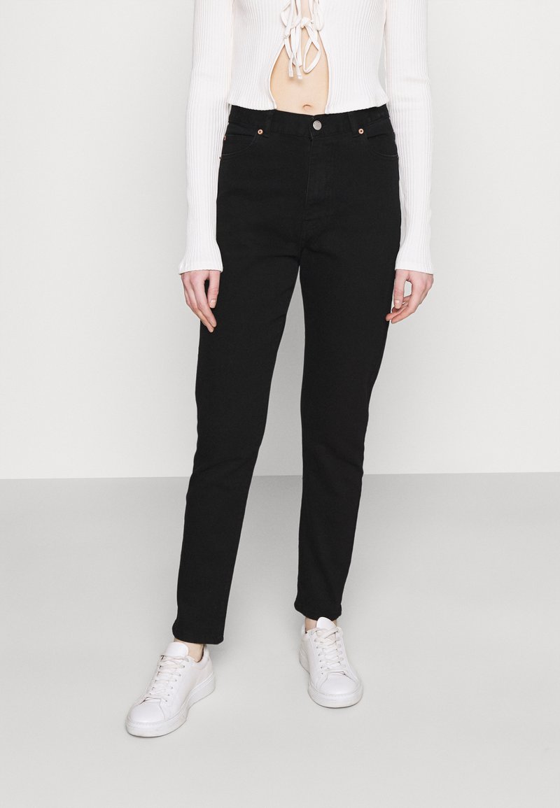 Jeans de denim preto com um corte slim, design de cinco bolsos e discretos detalhes em cobre. Combinados com uma blusa de manga longa branca canelada e ténis brancos.