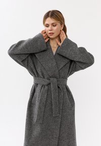 Cappotto lungo grigio in lana con maniche ampie e vita regolabile. Presenta una finitura testurizzata e un colletto profondo per ulteriore calore.