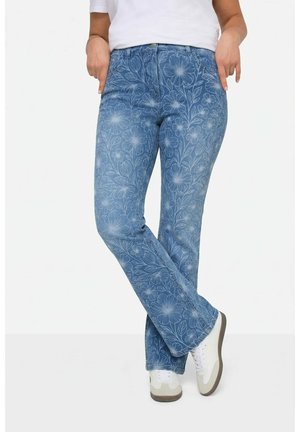 Femme portant un jean bleu à motif floral et des baskets blanches, debout les jambes croisées sur fond blanc.