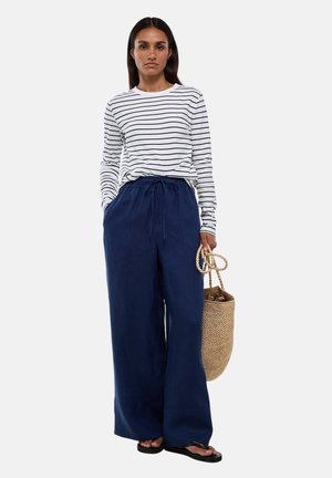 Femme portant une chemise blanche à manches longues avec des rayures noires, un pantalon large bleu marine, des tongs noires, tenant un sac en paille tressé à ses côtés.
