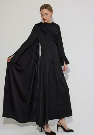 Kvinde iført en lang sort kjole med draperet stof på den ene skulder og udvidede ærmer, der holder kjolen ud, mens hun står mod en ensfarvet baggrund.