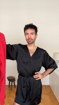 Svart satengkåpe med korte ermer og knytt liv; rødt mønstret plagg holdt i venstre hånd. Trekrakk i bakgrunnen.