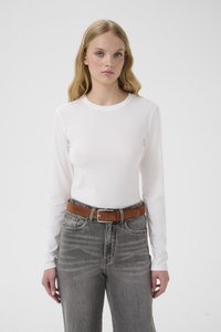 Ung kvinne med langt blondt hår iført en hvit langermet skjorte, grå høytlivsjeans og et brunt belte, stående foran en ensfarget bakgrunn.