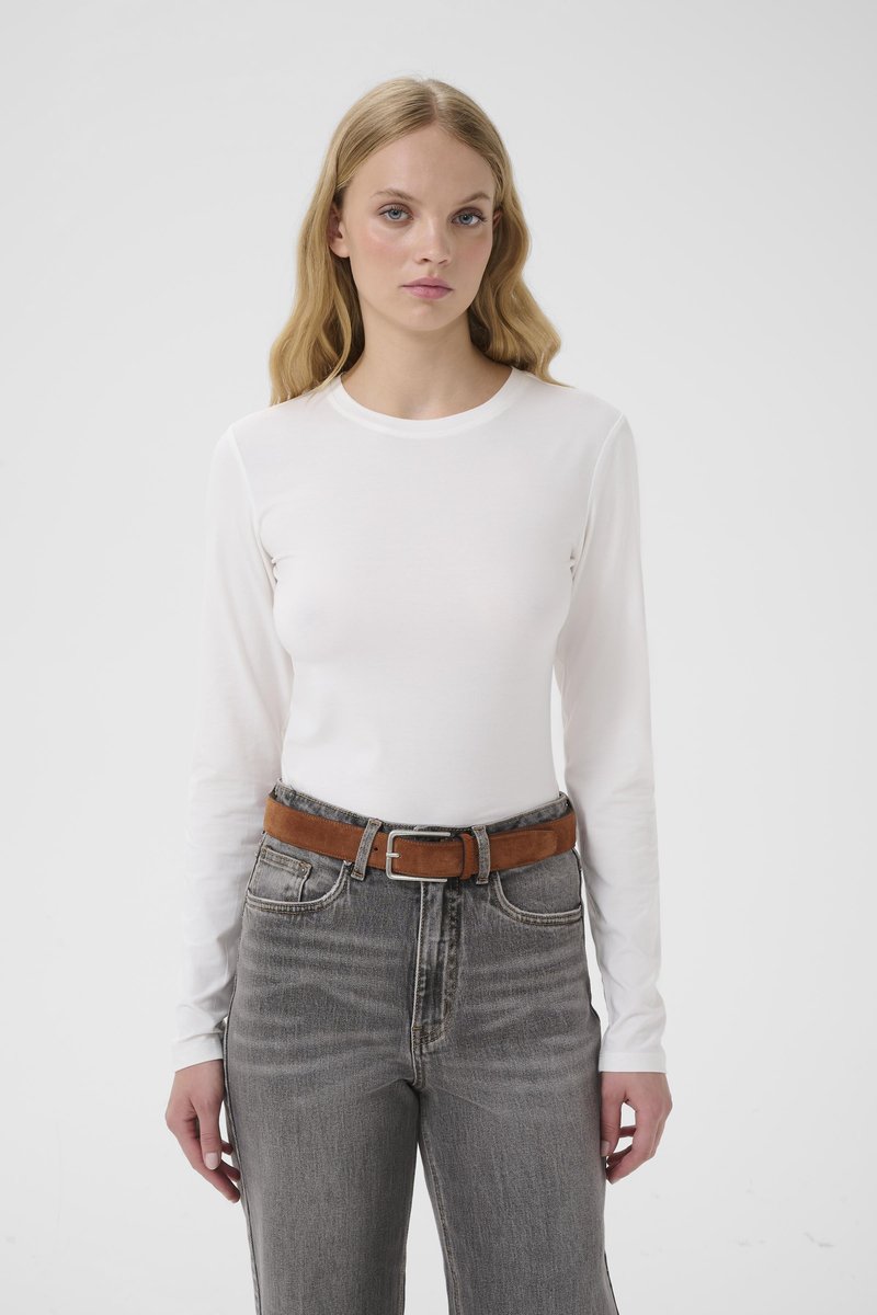 Ung kvinne med langt blondt hår iført en hvit langermet skjorte, grå høytlivsjeans og et brunt belte, stående foran en ensfarget bakgrunn.