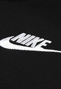 Juoda medžiaga su ryškiu baltu siuvinėtu Nike logotipu, demonstruojanti lygią tekstūrą ir aiškius, ryškius užrašus su lenktu akcentu.