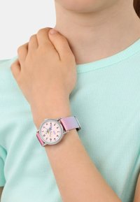 Ein Kind, das ein hellblaues Hemd trägt, zeigt eine Armbanduhr mit einem pink-lila glitzernden Armband und einer Feenillustration auf dem Zifferblatt.