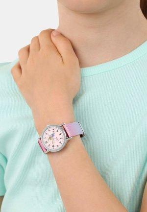 Ein Kind, das ein hellblaues Hemd trägt, zeigt eine Armbanduhr mit einem pink-lila glitzernden Armband und einer Feenillustration auf dem Zifferblatt.
