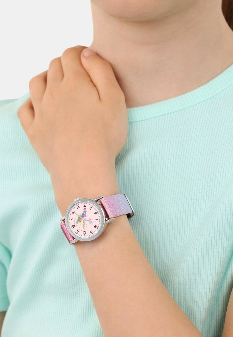 Ein Kind, das ein hellblaues Hemd trägt, zeigt eine Armbanduhr mit einem pink-lila glitzernden Armband und einer Feenillustration auf dem Zifferblatt.