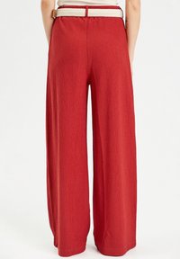 Pantalon rouge à jambes larges en tissu texturé, avec une ceinture tissée crème. La vue de dos montre une taille haute avec une ceinture lisse.