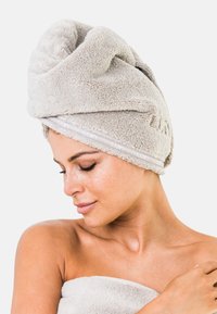 Toalla de pelo de microfibra gris, textura suave, diseño retorcido, con una banda interior de marca. La modelo la lleva envuelta en la parte superior de su cabeza.