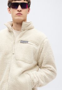 Μπεζ μπουφάν από φλις με ψηλό γιακά, φερμουάρ κλεισίματος και τσέπη στο στήθος. Υφή υφάσματος και εμφανές λογότυπο της μάρκας.