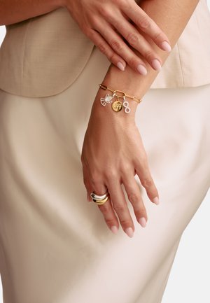 Gold chain bracelet with decorative charms: a bow, infinity symbol, and round medallion. Smooth links with a polished finish.