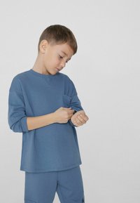 Modrá košile s dlouhým rukávem a kulatým výstřihem, se zipem na hrudi. Tkanina se zdá být měkká a texturovaná, v kombinaci s odpovídajícími modrými kalhotami.