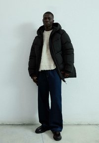 Svart puffjacka med huva, strukturerad tyg, beige stickad tröja under, lösa blå denimjeans och svarta skor.