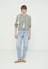 Cardigã cinza claro, jeans azul claro e sapatos bege com padrão. Modelo segura uma pequena bolsa branca. Roupa simples e casual.