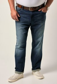 Mörkblå denimjeans med rak passform, subtil blekning och uppvikta fållar, kombinerat med vita sneakers. En brun läderbälte och klocka syns.