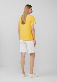 Gelbes T-Shirt mit kurzen Ärmeln, kombiniert mit weißen Shorts, die hochgekrempelt sind. Das Outfit wird mit beigen Turnschuhen abgerundet. Sauberes, schlichtes Design.