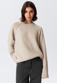 Vrouw met schouderlang donker haar, gekleed in een beige gebreide trui en donkergrijze jeans, staand tegen een effen lichte achtergrond.