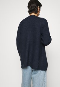 Granatowy dzianinowy sweter o luźnym kroju, z prążkowanymi mankietami i zaokrąglonym dekoltem, zestawiony z jasno-niebieskimi dżinsami.