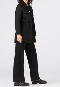 Svart skjortklänning med knappar, långa ärmar, midjebälte, två bröstfickor och vida svarta byxor. Slät tygstruktur och minimalistisk design.