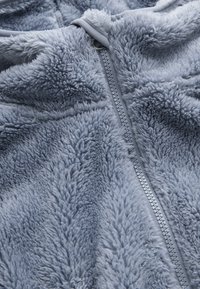 Helles blaues Fleece-Teil mit einer weichen, strukturierten Oberfläche und einem zentralen Reißverschluss. Der Stoff zeigt eine plüschige, flauschige Qualität.
