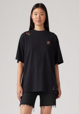 Femme aux longs cheveux noirs porte un T-shirt noir oversize avec une petite broderie florale et un short en jean noir aux ourlets effilochés.