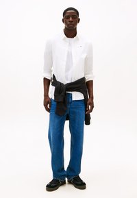 Camisa branca de botão por cima de uma t-shirt branca, combinada com jeans azuis. Um suéter escuro amarrado à cintura. Sapatos pretos, textura lisa.