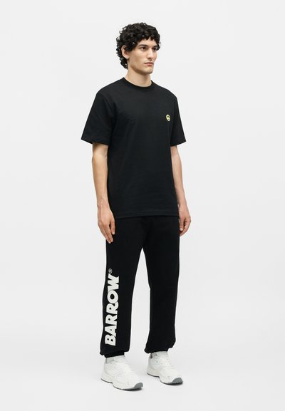Νέος άνδρας στέκεται φορώντας μαύρο μπλουζάκι με μικρό κίτρινο λογότυπο, μαύρο φόρμα με το κείμενο "BARROW" στο πόδι και λευκά αθλητικά παπούτσια.