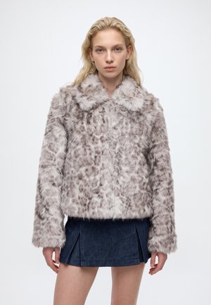 Un mannequin blond se tient devant un fond blanc, vêtu d’une veste grise à motifs en fausse fourrure et d’une jupe courte bleu foncé.