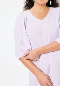 Helllilaes Kleid mit V-Ausschnitt und puffartigen Ärmeln, aus strukturiertem Stoff und mit taillierter Taille. Nahaufnahme von Arm und Hand.