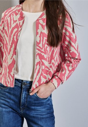 Mujer con cabello largo y oscuro que lleva una chaqueta con cremallera de patrón rosa y blanco sobre una camisa blanca y vaqueros azules, sobre un fondo gris.