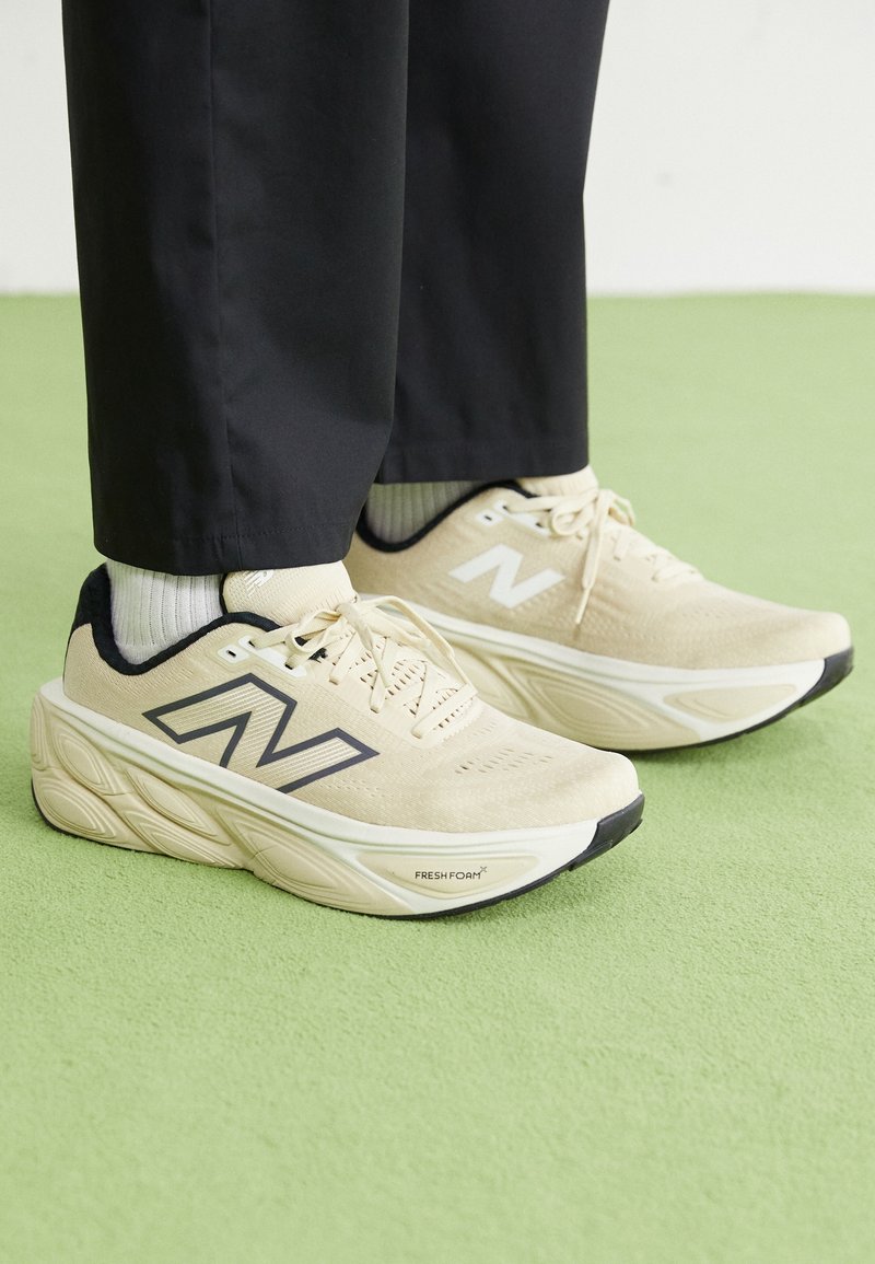 Baskets beige New Balance Fresh Foam portées avec des chaussettes blanches et un pantalon noir sur un sol vert.