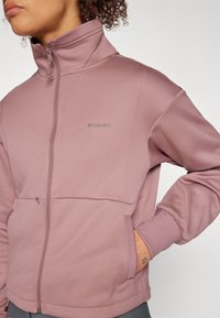 Fleece jacket in muted mauve with a high collar, zip front, and two side pockets. Features subtle brand logo and ribbed cuffs.