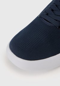 Navy blue sneaker with a textured knit upper, featuring a white rubber sole and traditional lacing system. Close-up of the toe area.