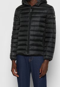 Sort oppustelig jakke med hætte, der har vandrette quiltede sektioner, lynlås foran og elastiske manchetter. Materialet ser skinnende og let ud.